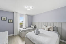 This serene bedroom, with its soft lavender walls and cozy upholstered beds, invites relaxation. Large windows let in natural light, enhancing the minimalist decor and comfortable textiles for a refreshing atmosphere.