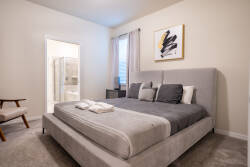 This elegantly designed bedroom invites relaxation with its plush gray bed and crisp white linens. Natural light filters through stylish curtains, enhancing the contemporary artwork and creating a soothing atmosphere for your.