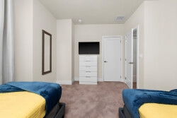 This bright, airy bedroom offers two cozy beds with rich blue throws, a sleek white dresser, and a wall-mounted TV for your entertainment, all set against a neutral palette and soft carpeting for a relaxing atmosphere.