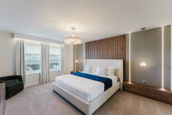 Enjoy the soft glow of the stylish chandelier and plush bedding in this contemporary bedroom. Large windows invite natural light, highlighting warm wooden accents for a cozy and refined atmosphere perfect for relaxation.