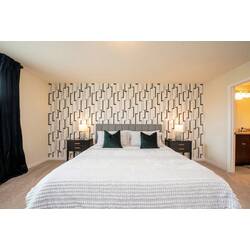 Rest in a spacious bedroom with a white textured bed, green accent pillows, geometric wall, black nightstands, soft lamps, dark curtains, and private bathroom access for vacation comfort.