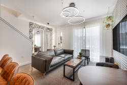 Enjoy the warmth of natural light pouring through large windows, highlighting the sleek leather sofa and textured decor, as a modern chandelier creates an inviting atmosphere perfect for relaxation and great conversations.