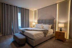 Enjoy ultimate relaxation in this stylish bedroom featuring a plush oversized bed with crisp white linens. Soft gray curtains frame large windows, and gentle lighting creates a cozy ambiance for restful nights.