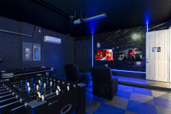 Welcome to our lively game room, where fun awaits with a foosball table and a large screen for movies. The vibrant blue lighting against the sleek black walls sets the perfect mood for relaxation and entertainment.