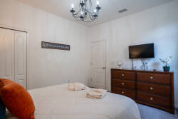This cozy bedroom is bright and airy, featuring a plush bed with soft towels and a stylish orange accent pillow. A charming chandelier provides a warm glow, and the sleek dresser adds ample storage for a relaxing retreat.