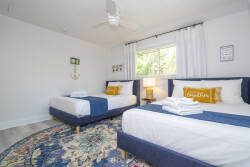 Experience a bright and airy bedroom with two cozy beds dressed in crisp white linens and vibrant blue accents. A charming rug adds warmth, while natural light pours in through the large window, creating a welcoming ambiance.