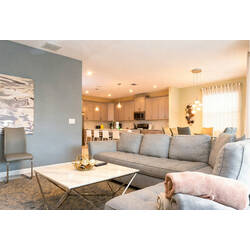 Relax in a bright open living area with a spacious gray sectional, marble-style coffee table, soft curtains, warm lighting, modern decor, and open access to the kitchen and dining space.