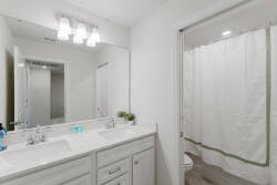 Experience a bright and clean spacious bathroom with dual sinks, elegant chrome fixtures, and a soothing palette of whites and soft greens. Ample lighting and a modern shower curtain enhance your daily routines for pure enjoyment.