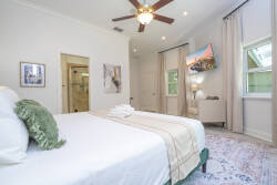 This cozy bedroom boasts a plush white bed with soft linens and a decorative green pillow, creating a warm atmosphere. Natural light pours in through large windows, enhancing the serene vibe alongside a stylish ceiling fan.