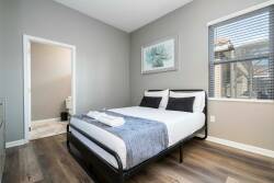 Natural light streams into the cozy bedroom, highlighting soft gray walls and stylish wood flooring. The neatly made bed with crisp white linens and a textured throw invites relaxation, complemented by charming artwork.