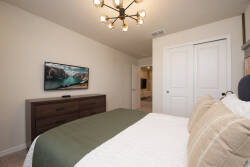 Enjoy comfort in this cozy bedroom with a plush bed dressed in soft linens and a stylish green accent. A rustic chandelier brightens the room, while a sleek dresser and wall-mounted TV provide modern convenience.