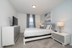 Enjoy a soothing atmosphere in this stylish bedroom with plush light gray carpeting and calming blue walls. The inviting queen bed and modern furnishings promise restful nights, complemented by a sleek dresser and wall-mounted TV.