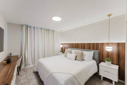 This cozy bedroom boasts a plush queen bed with soft neutral bedding, a warm wooden accent wall, and natural light flowing through elegant curtains, all enhanced by stylish pendant lighting for a serene atmosphere.