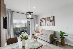 Natural light streams through large windows, illuminating the cozy beige sofa and modern dining area adorned with sleek tableware. A touch of greenery adds freshness, while artistic wall decor enhances the inviting atmosphere.