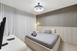 Experience a cozy bedroom with a stylish upholstered bed in soft gray linens, enhanced by textured wall panels. Warm light from a modern ceiling fixture creates a serene atmosphere, perfect for restful nights.