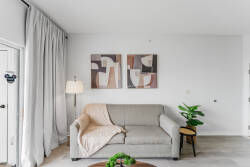 Unwind on the soft gray sofa with a cozy blanket, surrounded by two abstract artworks that bring creativity to the airy space. A stylish lamp and a potted plant add to the inviting decor, making your stay truly welcoming.