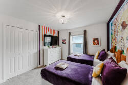 This bright bedroom features two cozy twin beds with soft purple bedding, vibrant artwork, and playful decor. Enjoy ample storage with a sleek dresser and built-in closets, all bathed in warm natural light for a welcoming feel.