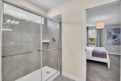 Experience a bright, modern bathroom with sleek glass shower doors and elegant gray tiling, creating a clean, inviting space. Beyond, a softly lit bedroom with a cozy purple throw beckons you to relax and unwind in tranquility.