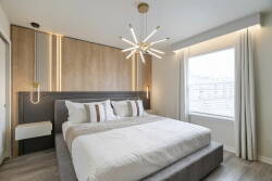 This stylish bedroom offers a plush king-size bed with crisp linens and decorative pillows, warm wood paneling, and a unique starburst chandelier that casts a soft glow, all enhanced by natural light from the expansive window.