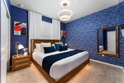 This stylish bedroom features a striking blue accent wall that beautifully contrasts with soft neutral bedding and carpet. Enjoy the ambient glow of a modern light fixture, paired with warm, sleek wooden furniture for a cozy feel.