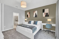 Experience comfort and style in this chic bedroom, featuring a plush gray carpet, a sleek white bed with textured throw pillows, inviting soft lighting from bedside lamps, and sophisticated wall art that enhances the ambiance.