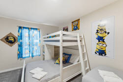 This bright children's room features a cozy white bunk bed with soft gray bedding, colorful Minions decor, and vibrant blue curtains, creating a playful and delightful space for kids to relax and enjoy their time.