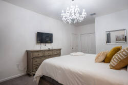 Experience a bright and airy bedroom with a plush bed featuring soft pillows and an elegant chandelier. The warm wood dresser complements the serene white walls, creating a cozy and inviting atmosphere for your stay.