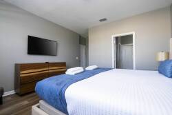 Unwind in this cozy bedroom with a plush king-size bed draped in crisp white linens and a rich blue accent blanket. The warm wood dresser pairs perfectly with the sleek wall-mounted TV, all bathed in natural light.