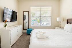 Bright and airy, this cozy bedroom features a plush white bed adorned with neatly arranged towels, a sleek dresser, and a large wall-mounted TV. Sunlight filters through the blinds, creating a warm ambiance perfect for relaxation.