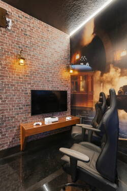 Experience a chic gaming room with a stunning exposed brick wall and warm sconces for a cozy vibe. Featuring sleek black gaming chairs and a modern entertainment setup, it’s the ideal spot for relaxation and excitement.