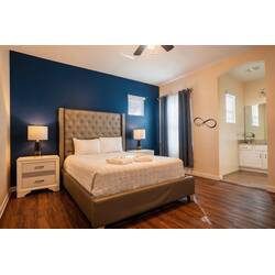 Experience comfort in this cozy bedroom with a plush tufted headboard against a deep blue wall, natural light pouring in through large windows, warm wood flooring, stylish decor, and an en-suite bathroom for your privacy.