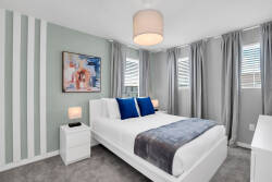 Bathed in natural light, this cozy bedroom features a crisp white bed adorned with a soft gray throw and vibrant blue pillows, complemented by modern artwork and soothing striped walls that enhance the tranquil atmosphere.
