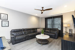 Relax in this spacious living room featuring a sleek leather sectional that invites conversation. The subtle gray walls complement the modern decor, while the fan overhead and natural light create a warm, welcoming ambiance.