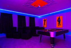 Step into a vibrant entertainment room featuring a sleek pool table, stylish black seating, and captivating pop art on the walls. The bold purple and blue lighting sets a dynamic atmosphere, perfect for gathering with friends.