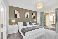 This stylish bedroom boasts a plush king-size bed with crisp linens and accent pillows, complemented by soft gray carpeting. Large windows with elegant drapes invite natural light, highlighting the modern artwork on the walls.