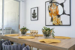 The dining area features a beautifully set wooden table with vibrant yellow placemats and elegant glass centerpieces, while colorful abstract artwork on the walls creates an inviting and warm atmosphere for guests.