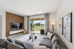 Relax in a bright living room with cozy gray seating, a wall-mounted TV, warm wood accent wall, soft curtains, modern decor, and sliding glass doors opening to a peaceful balcony view.