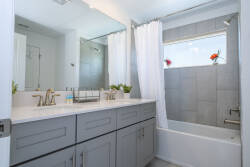 This bright bathroom showcases a sleek gray vanity with plenty of storage and stylish gold fixtures. A large mirror reflects natural light from the window, creating a fresh atmosphere ideal for a refreshing start to your day.