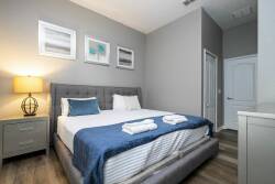 Enjoy restful nights in this beautifully designed bedroom with a plush gray upholstered bed, crisp white linens, and soft blue accents. The warm bedside lamp and subtle artwork offer a soothing ambiance for ultimate relaxation.