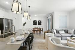 Discover our airy open-concept space that seamlessly blends the kitchen and living area, showcasing modern cabinetry and a chic gray sectional, all illuminated by pendant lights for a warm ambiance ideal for meals and laughter.