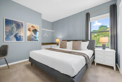 Relax in this serene bedroom featuring soothing blue walls, fresh white bedding, and tasteful artwork. Enjoy the natural light pouring in through the large window, offering a lovely view of greenery and the warm glow of stylish.