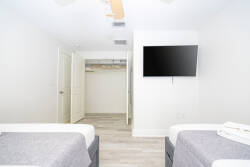 Step into a bright and airy bedroom featuring two cozy beds adorned with soft gray linens, perfect for a restful night's sleep. A sleek TV on the wall adds a modern touch, while the light wood flooring enhances the spacious feel.