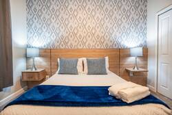 Rest in a cozy bedroom with a clean white bed, blue accent throw, wood headboard, geometric feature wall, soft lamps, gray curtains, and warm natural details, creating a comfortable space for vacation.