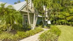 Step into a vibrant landscape lined with lush greenery and palm trees, perfectly framing the charming entrance of your vacation home. The welcoming path leads you to a serene setting where nature and comfort harmoniously coexist.