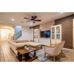 Relax in a warm and elegant living room with a spacious sectional sofa, wall-mounted TV, white cabinets, and soft natural light through the curtains. Neutral tones, wood floors, and cozy seating create a welcoming space for family time.