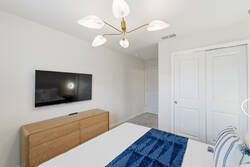 Rest in a bright modern bedroom with a cozy white bed, blue throw, wood accent wall, soft pendant lights, white curtains, wall-mounted TV, and calm neutral details for vacation comfort.