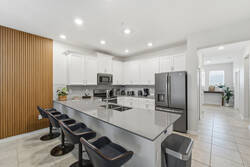 Cook with ease in a bright modern kitchen with white cabinetry, stainless steel appliances, a large island, bar seating, warm wood accents, and an open view toward the entryway.