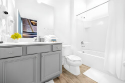 Enter this bright bathroom with a sleek gray vanity against crisp white walls. Warm wood flooring adds comfort, and well-placed lighting enhances the airy atmosphere, creating a refreshing space for you to relax and unwind.