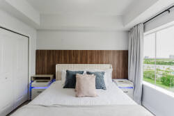 Awaken in a cozy bedroom with a plush white bed, textured pillows, and calming blue tones. Large windows let in natural light and showcase a refreshing view of greenery, while warm wooden paneling adds a comforting touch.