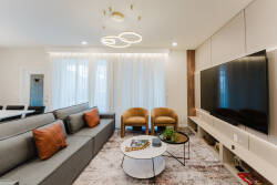 Enjoy the bright and inviting living room with a plush gray sofa adorned with vibrant orange pillows. The modern chandelier and sleek media console, along with a stylish coffee table, create a comfortable space for your.